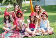 Two women and four girls enjoy a sunny picnic in Portugal, waving happily in the park.