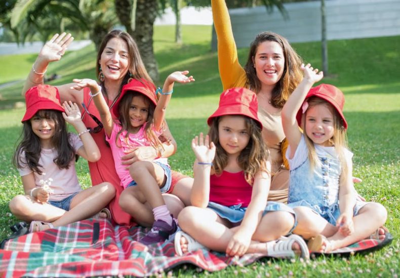 Two women and four girls enjoy a sunny picnic in Portugal, waving happily in the park.